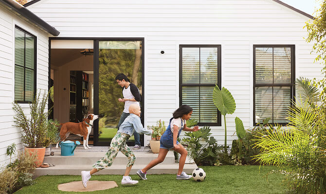 Two children play soccer in a backyard while an adult and a dog stand near an open door to the house. A Google Nest Cam (outdoor or indoor, battery) is installed on the side of the house.